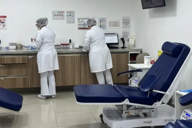 Duas profissionais de saúde de costas em uma sala de coleta, balcão com materiais e poltronas reclináveis azuis