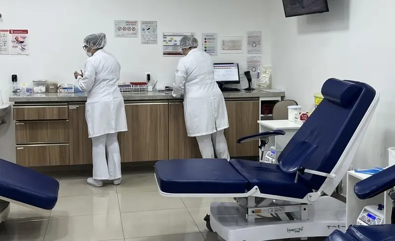 Duas profissionais de saúde de costas em uma sala de coleta, balcão com materiais e poltronas reclináveis azuis