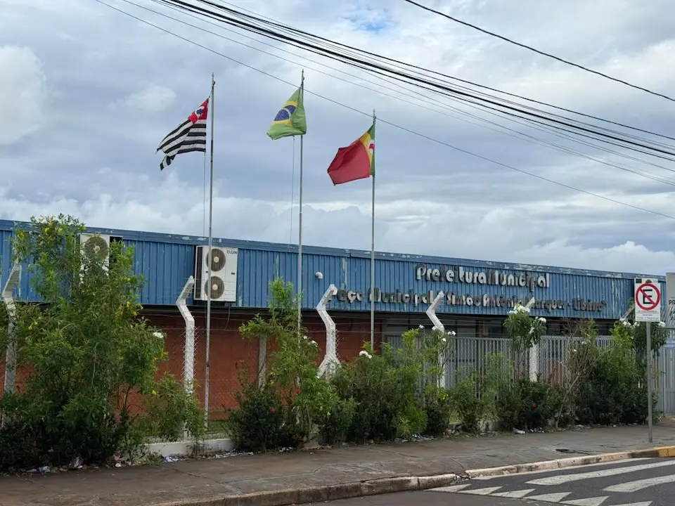 Fachada da Prefeitura Municipal de Barretos com três mastros de bandeiras, cerca e calçada em primeiro plano