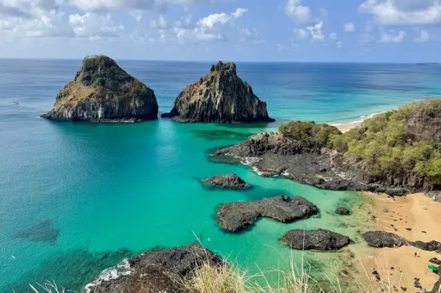 Baía com água turquesa, duas ilhotas rochosas e praia de areia com vegetação