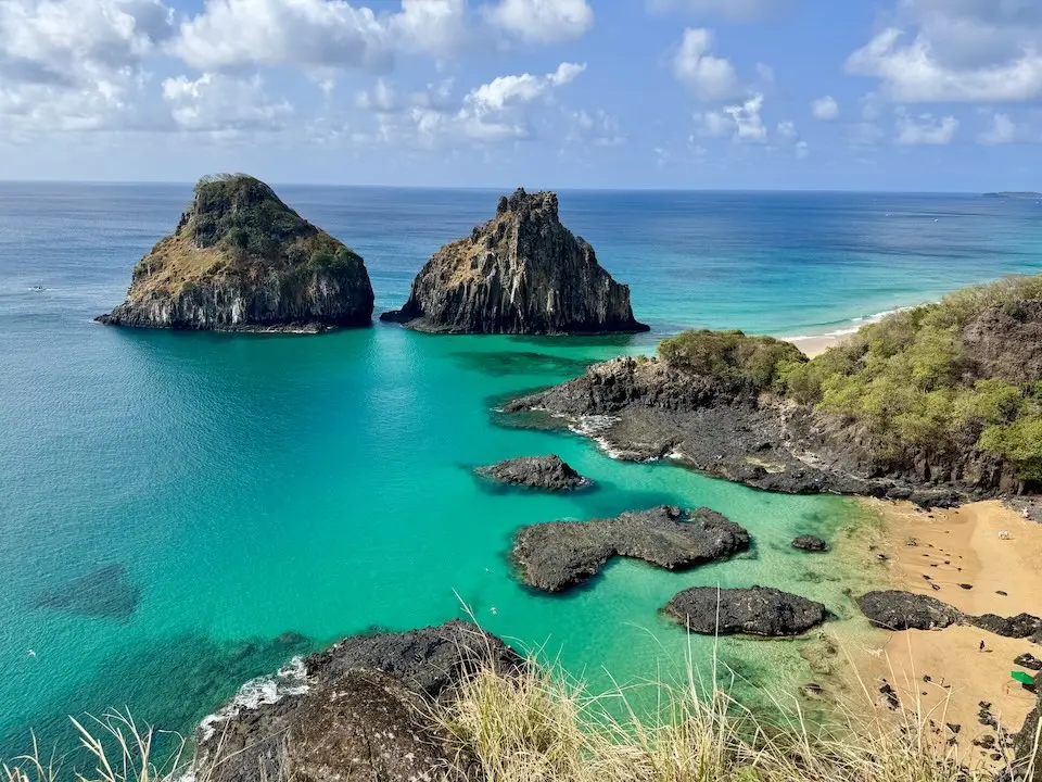 Baía com água turquesa, duas ilhotas rochosas e praia de areia com vegetação