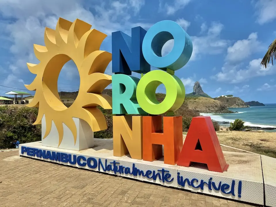 Letreiro colorido com as letras 'NORONHA' e um sol estilizado, vista de costa rochosa e mar ao fundo