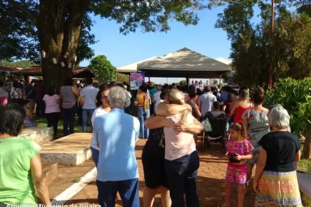 Pessoas reunidas em cerimônia ao ar livre no cemitério, com duas mulheres se abraçando em primeiro plano e uma tenda onde ocorre a celebração ao fundo