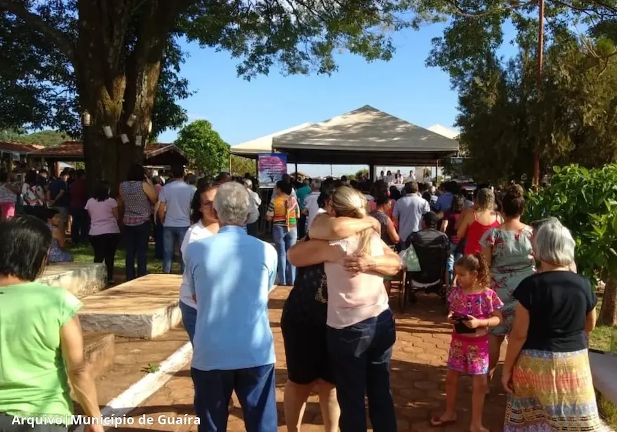 Pessoas reunidas em cerimônia ao ar livre no cemitério, com duas mulheres se abraçando em primeiro plano e uma tenda onde ocorre a celebração ao fundo