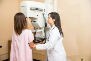 paciente posicionada em aparelho de mamografia enquanto profissional de saúde de jaleco ajusta o equipamento