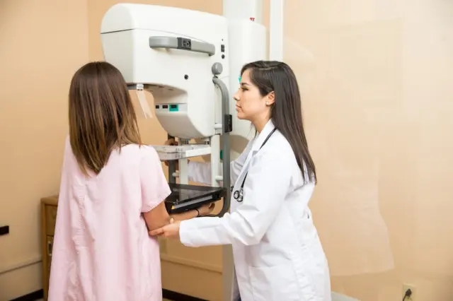 paciente posicionada em aparelho de mamografia enquanto profissional de saúde de jaleco ajusta o equipamento