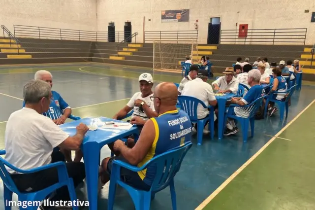 Pessoas idosas sentadas em mesas plásticas azuis disputando jogos de cartas e dominó em quadra coberta