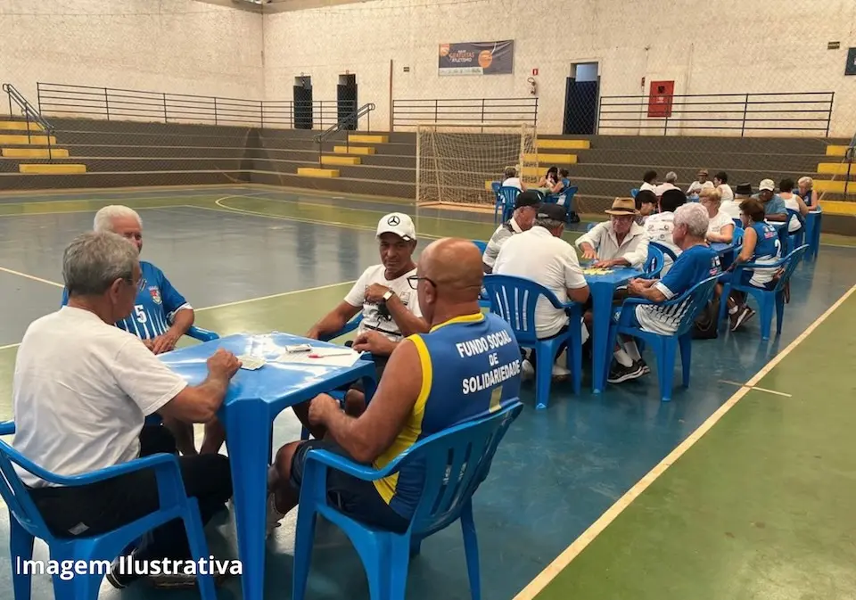 Pessoas idosas sentadas em mesas plásticas azuis disputando jogos de cartas e dominó em quadra coberta