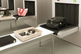 Esteira de inspeção em aeroporto com mala de mão preta sobre a esteira e bandeja plástica contendo fones de ouvido, carteira e relógio; ao fundo, botas e uma sacola listrada vermelha e branca