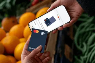 Duas mãos segurando um smartphone com imagem de um cartão na tela e um cartão físico azul da RecargaPay, com fundo de laranjas e folhagens