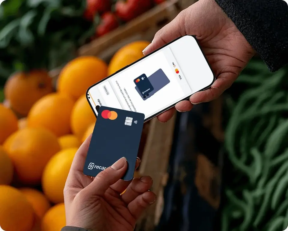 Duas mãos segurando um smartphone com imagem de um cartão na tela e um cartão físico azul da RecargaPay, com fundo de laranjas e folhagens