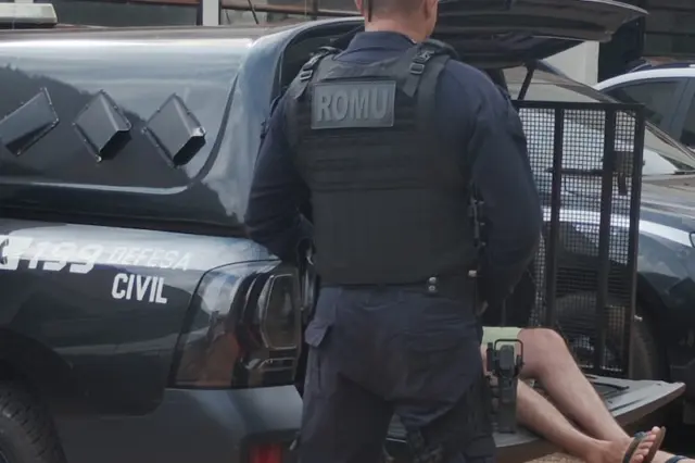 Agente da ROMU de costas ao lado de viatura com inscrição 'DEFESA CIVIL' e homem com as pernas para fora do compartimento traseiro