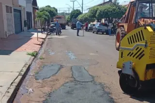 Rua residencial com trechos de asfalto fresco no centro, rolo compressor amarelo à direita e operários ao fundo