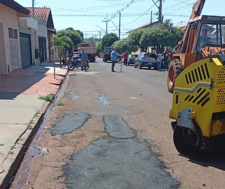 Rua residencial com trechos de asfalto fresco no centro, rolo compressor amarelo à direita e operários ao fundo