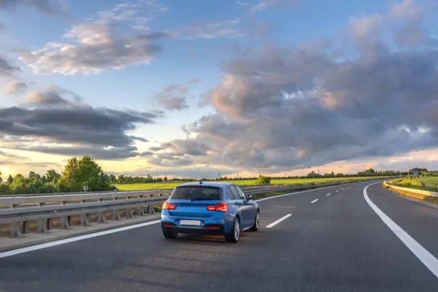 Carro hatchback azul circulando por uma rodovia com duas faixas, guarda-corpo metálico à esquerda e céu com nuvens ao entardecer